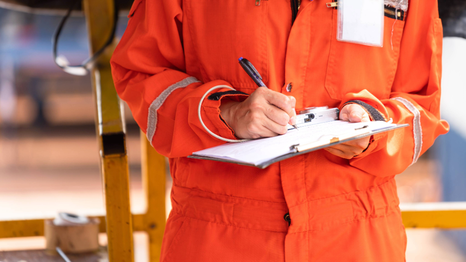 Arbeiter in einem leuchtend orangefarbenen Sicherheitsanzug schreibt mit einem Stift auf einem Klemmbrett neben einem gelben Geländer auf einem Industriegelände.
