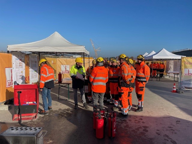 Arbeitssicherheitstage_MaxBögl_1 Mehrere Bauarbeiter in orangefarbenen Westen und gelben Helmen nehmen an einer Sicherheitsunterweisung in der Nähe von Zelten teil, mit roten Feuerlöschern.