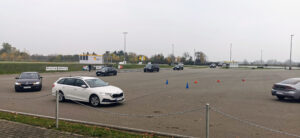Bedecktes Fahrevent auf einem Parkplatz mit orangefarbenen und blauen Kegeln; weißer Kontrollstand, Banner und mehrere Autos, Bäume im Hintergrund.