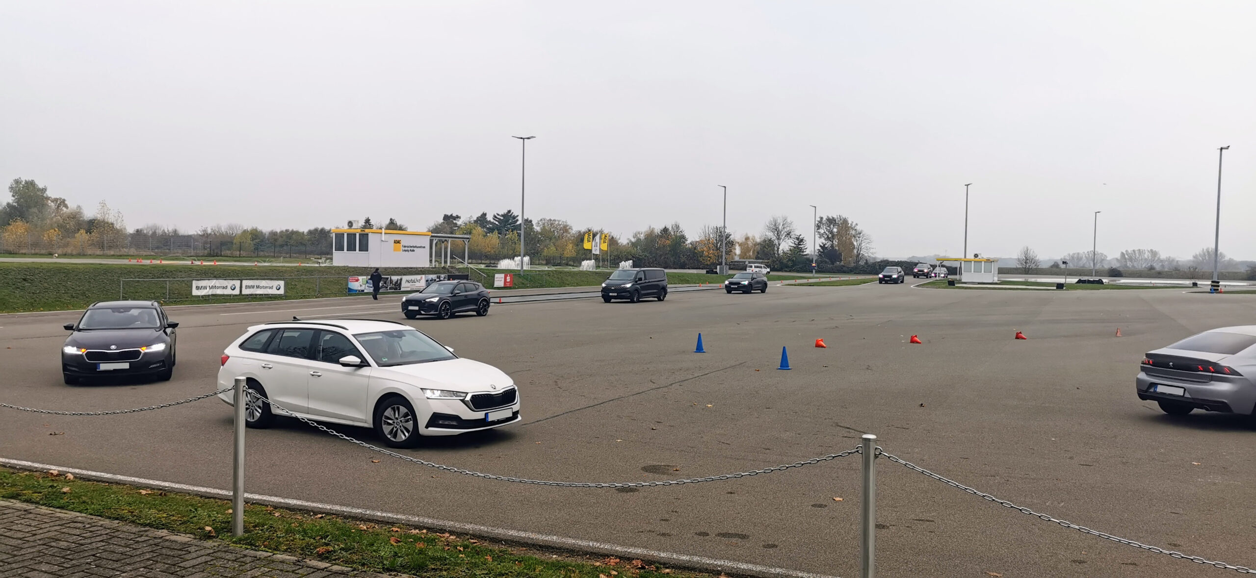 Bedecktes Fahrevent auf einem Parkplatz mit orangefarbenen und blauen Kegeln; weißer Kontrollstand, Banner und mehrere Autos, Bäume im Hintergrund.