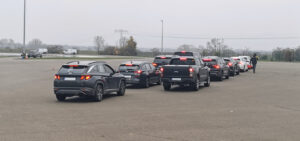Reihe von grauen und schwarzen SUVs und Pickups auf einem geräumigen, leeren Parkplatz an einem bewölkten Tag; eine Person mit einer roten Tasche geht entlang.