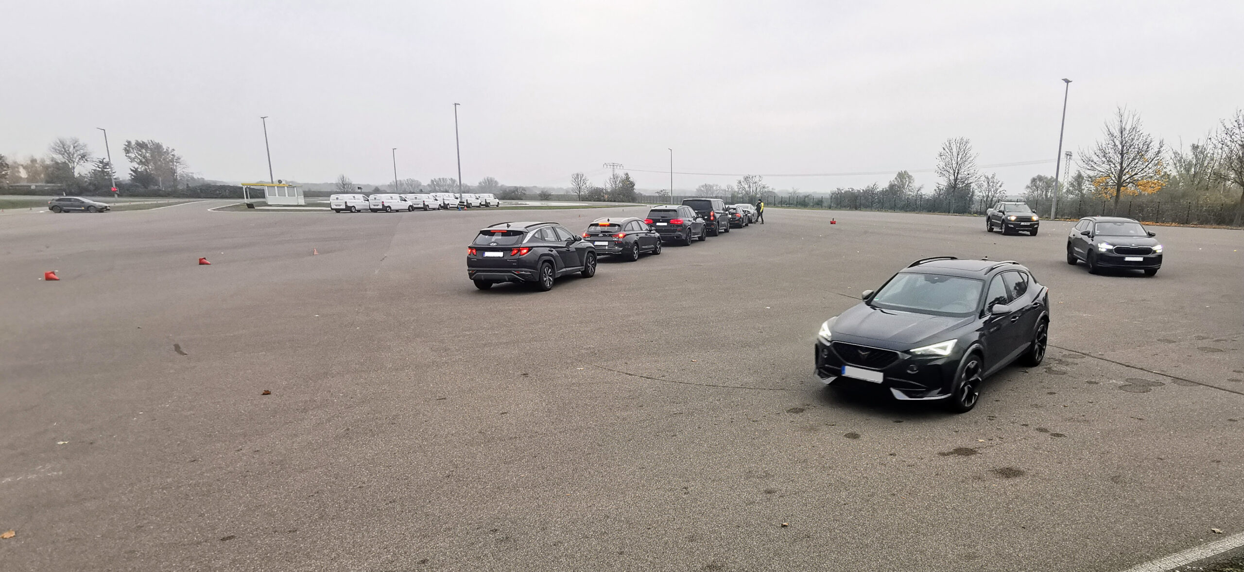 Reihe von Autos auf einem großen leeren Parkplatz, mit einem grauen SUV im Vordergrund; orangefarbene Kegel markieren das Gebiet, und eine kleine Hütte mit gelbem Dach steht weiter hinten.