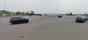 Weitläufiger Parkplatz mit orangenen Kegeln, vorne ein blauer Kegel; ein schwarzer SUV fährt vorbei, während weiße Vans sich in der Nähe eines kleinen Gebäudes aufstellen.