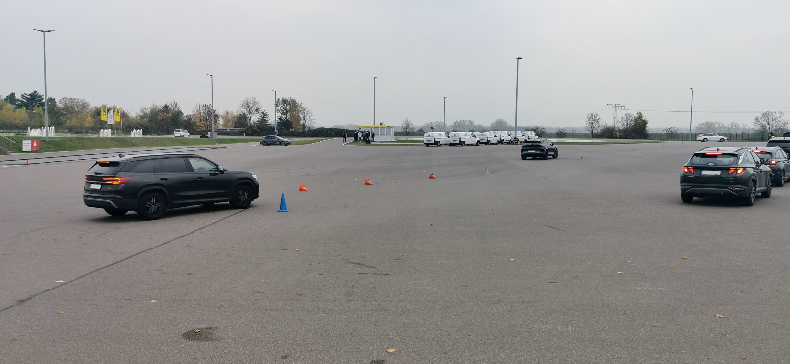 Weitläufiger Parkplatz mit orangenen Kegeln, vorne ein blauer Kegel; ein schwarzer SUV fährt vorbei, während weiße Vans sich in der Nähe eines kleinen Gebäudes aufstellen.
