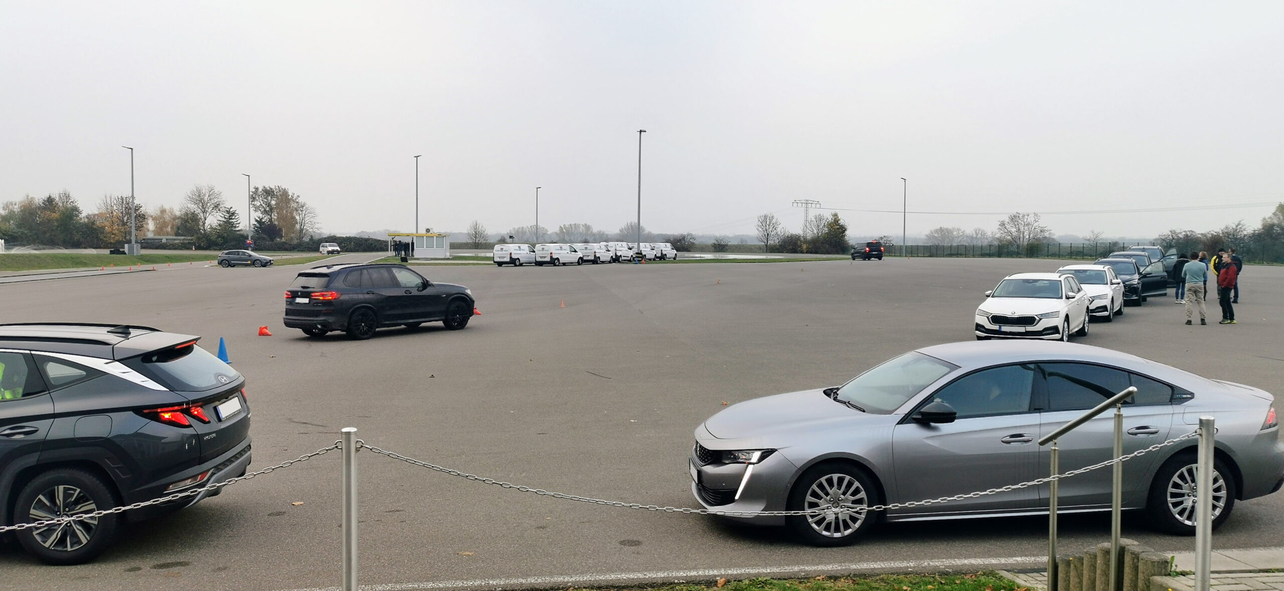 Bewölkter Parkplatz: Schwarzer SUV schlängelt sich um orangefarbene Kegel in der Nähe einer Reihe weißer Lieferwagen; einige Leute stehen an einem gelben Stand.