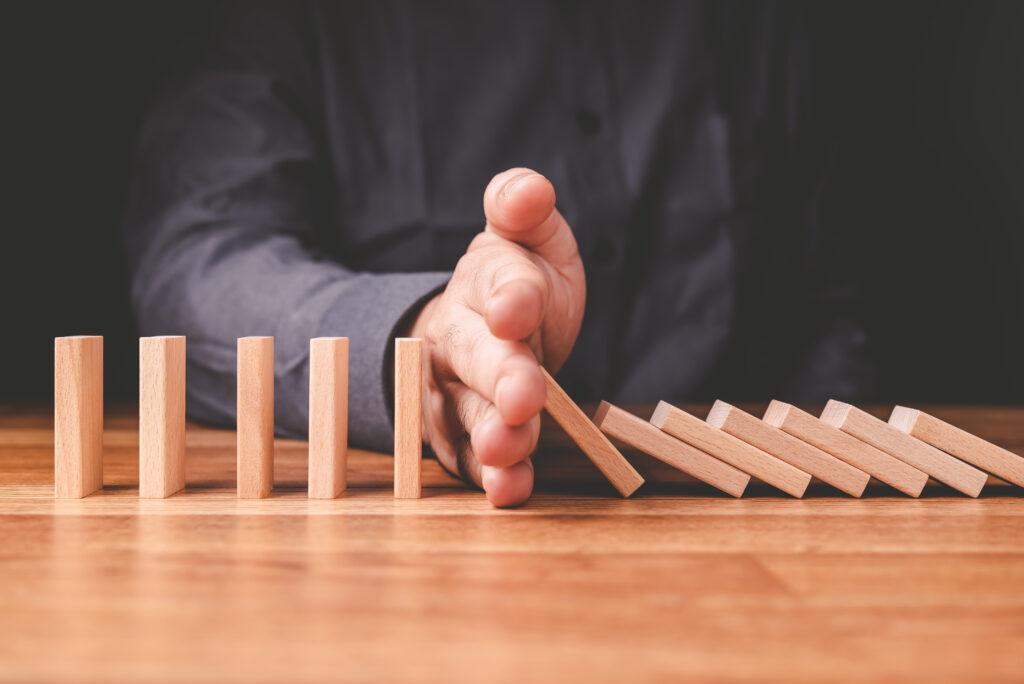 Nahaufnahme einer Hand, die eine Reihe von Holz-Domino-Steinen auf einem Holztisch anhält; aufrecht stehende Steine bleiben stehen, während andere umfallen.