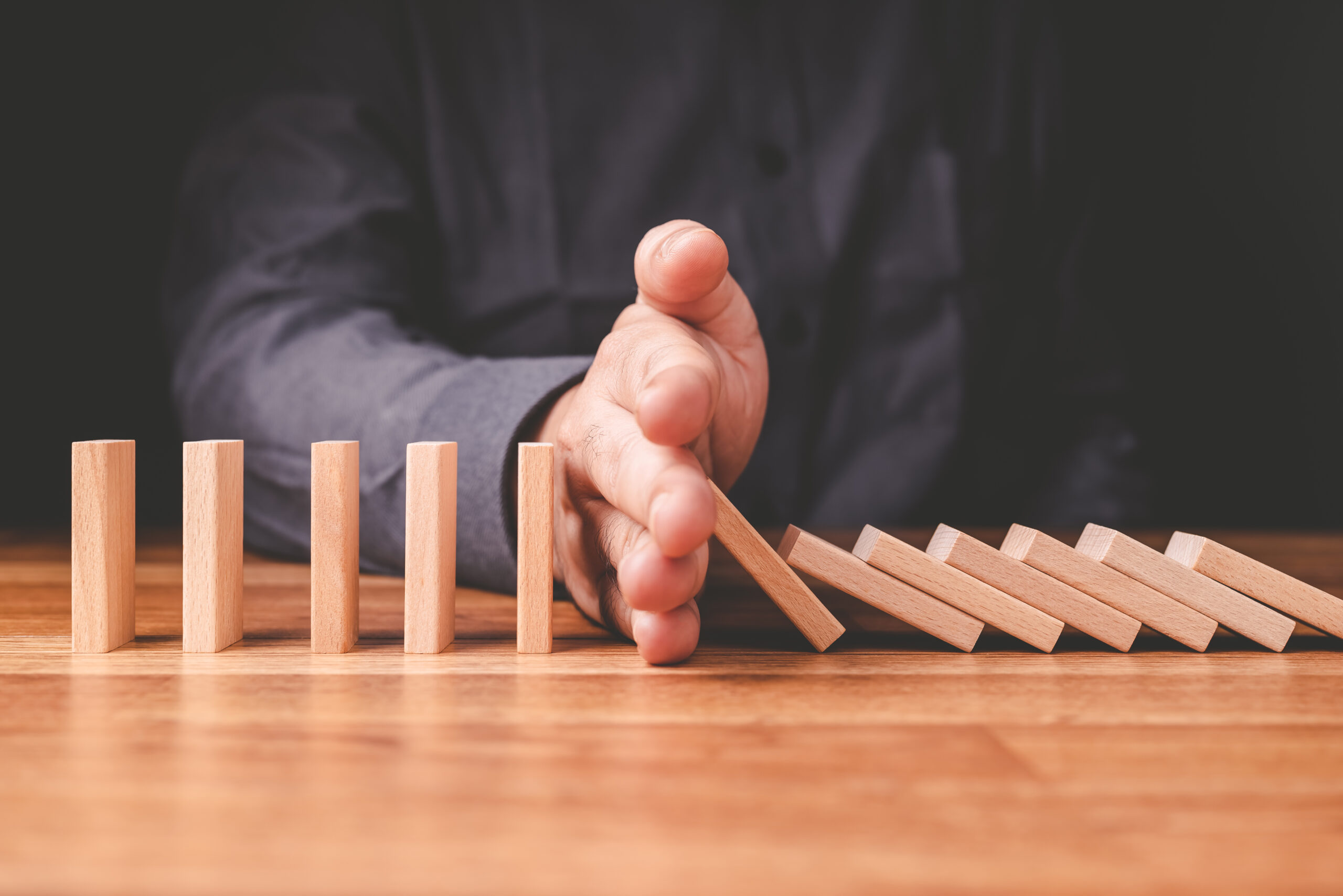 Nahaufnahme einer Hand, die eine Reihe von Holz-Domino-Steinen auf einem Holztisch anhält; aufrecht stehende Steine bleiben stehen, während andere umfallen.