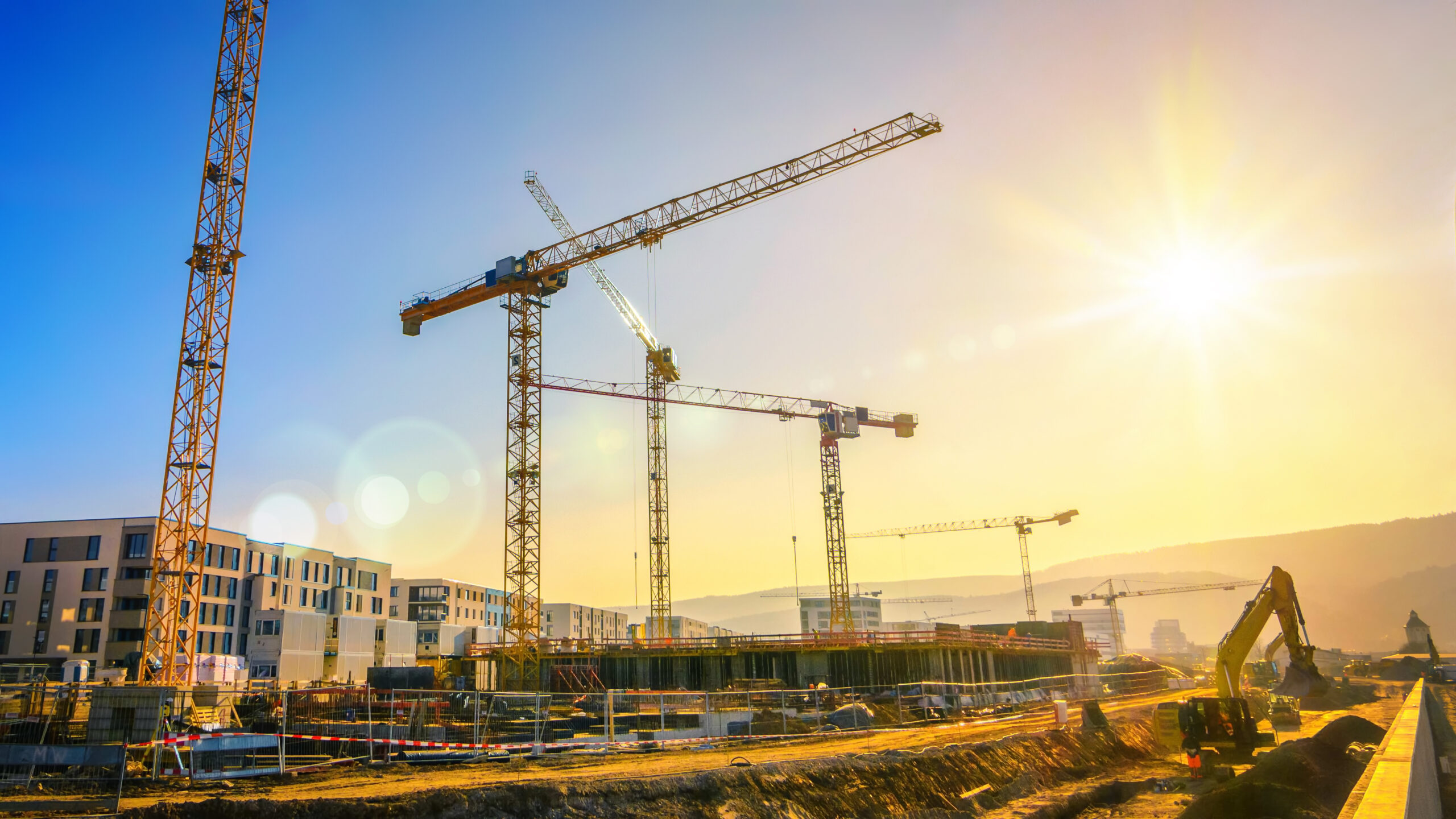 Sonnendurchflutete Baustelle mit hohen gelben Kränen über modernen weißen Wohnblöcken, blauer Himmel und Sonnenschein.