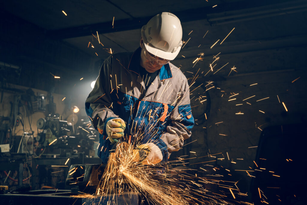 Arbeiter mit weißem Sicherheitshelm und Schutzbrille schleift Metall auf einer Werkbank, während helle Funken in einer dunklen Industriehalle fliegen.