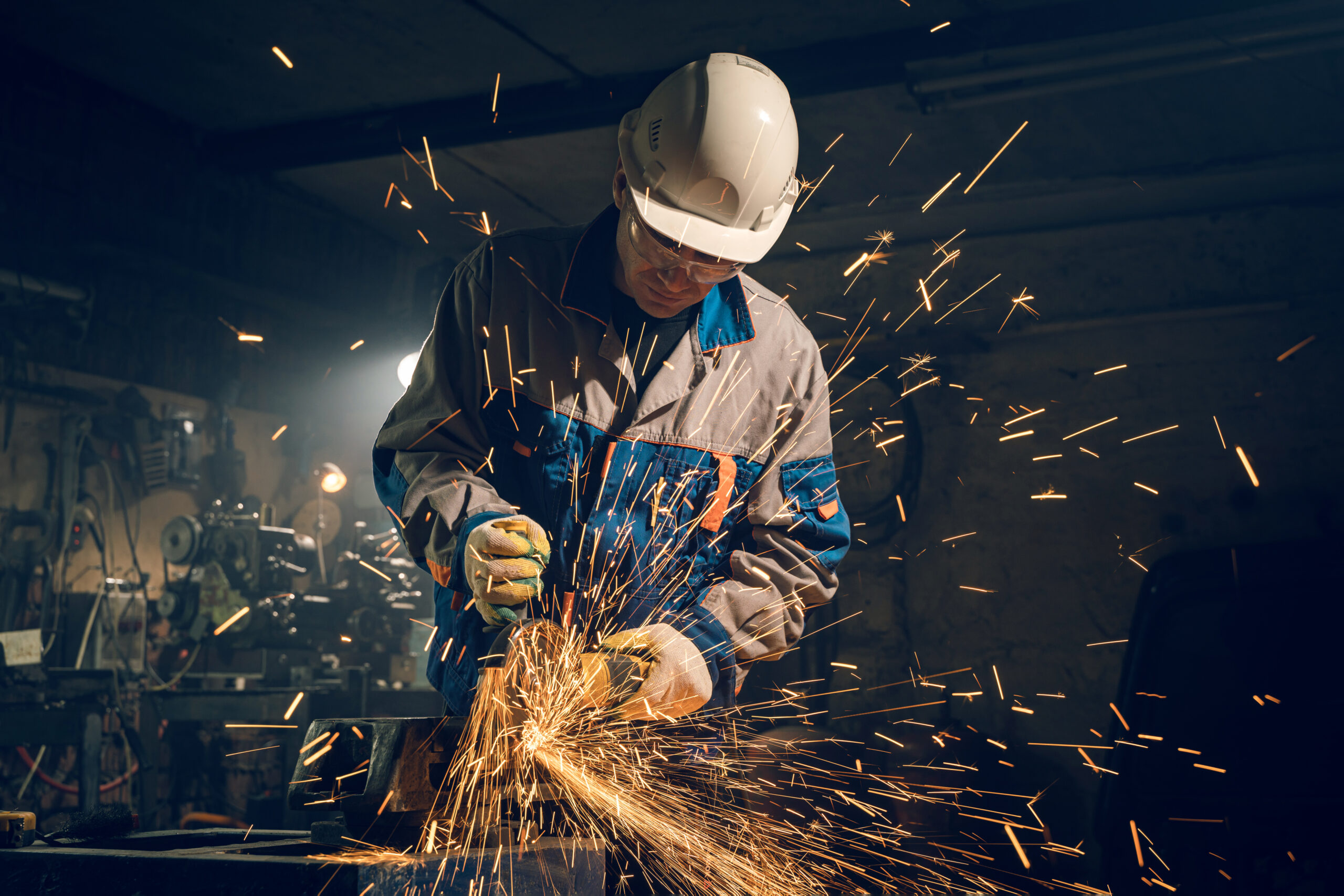Arbeiter mit weißem Sicherheitshelm und Schutzbrille schleift Metall auf einer Werkbank, während helle Funken in einer dunklen Industriehalle fliegen.