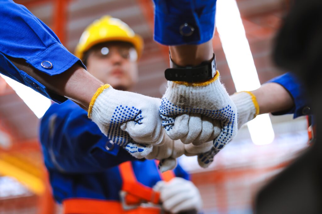 Nahaufnahme von behandschuhten Händen, die sich zu einem Kreis zusammenschließen, Arbeiter in blauen Uniformen mit einem gelben Schutzhelm vor einer Backsteinwand sichtbar.