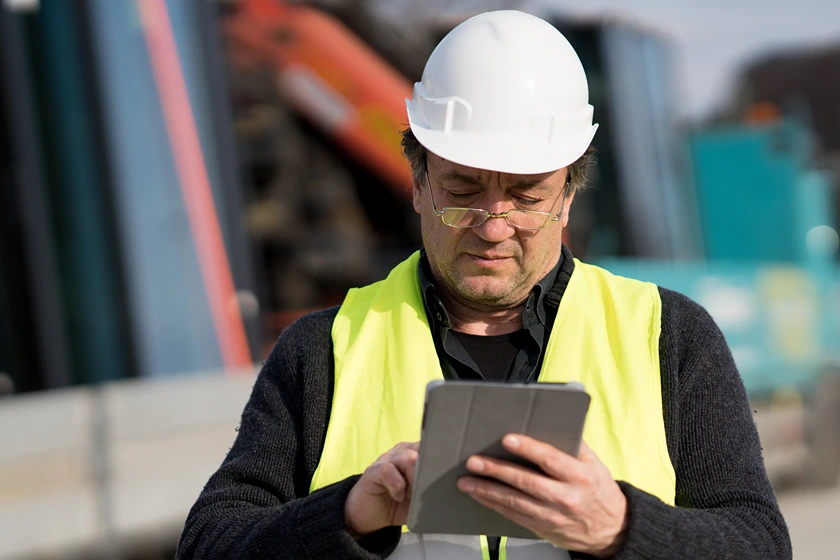 Ein Bauarbeiter mit weißem Schutzhelm und neongelber Weste benutzt ein Tablet auf einer Baustelle; im Hintergrund sind Maschinen verschwommen zu sehen.