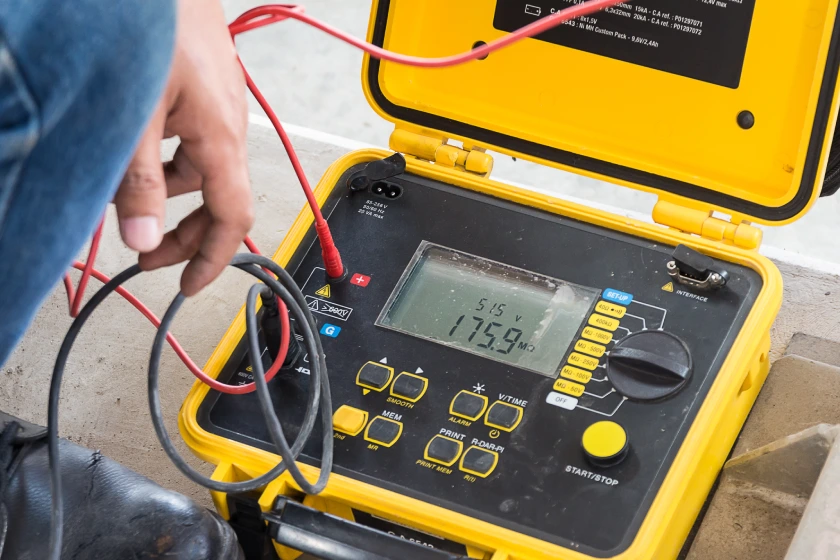 bb_Prüfungen-elektrischer-Betriebsmittel Techniker verwendet rote und schwarze Prüfspitzen an einem gelben robusten digitalen Elektrometer, Deckel offen, auf Beton.