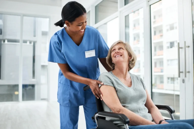 Eine Pflegekraft in blauer Schutzkleidung schiebt einen Rollstuhl, während eine ältere Frau in Grau in einer hellen Klinik lächelt.