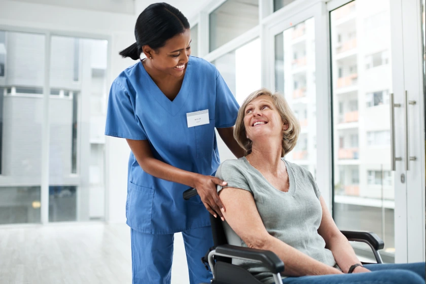 Eine Pflegekraft in blauer Schutzkleidung schiebt einen Rollstuhl, während eine ältere Frau in Grau in einer hellen Klinik lächelt.