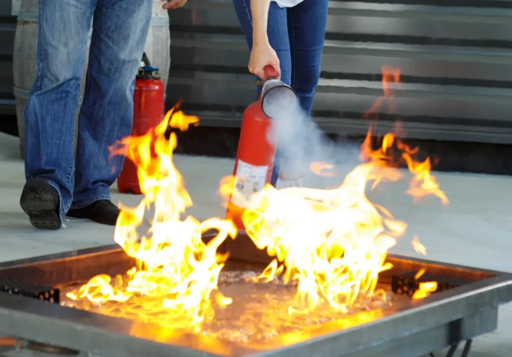 lp-brandschutzhelferausbildung-loeschen Brandschutzhelferausbildung: Übungsszene, zwei Personen beim Löschen eines Feuers mit Feuerlöscher, Flammen und Rauch.