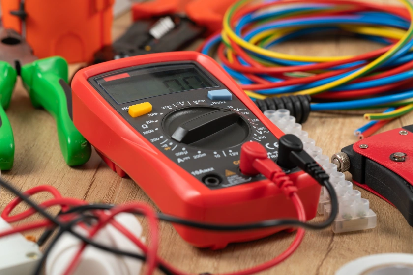 lp-elektr_pruefgeraet Roter Digital-Multimeter mit gelbem Knopf, Messleitungen und grünen Klemmen, farbige Kabel.